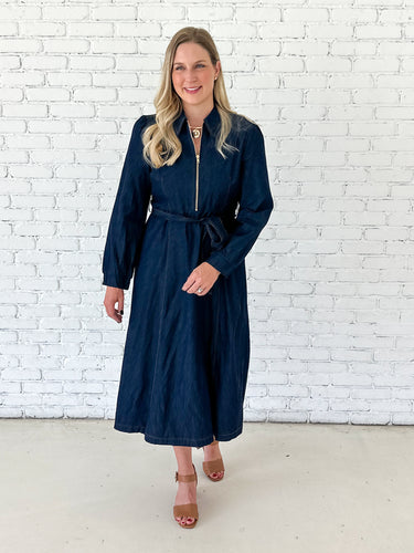A woman modeling a dark navy blue midi dress with a front zipper, tie waist belt, and long sleeves, standing against a white brick wall.
