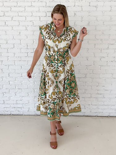 A woman modeling a sleeveless midi dress with a green and gold baroque-style pattern on a cream background, standing against a white brick wall.