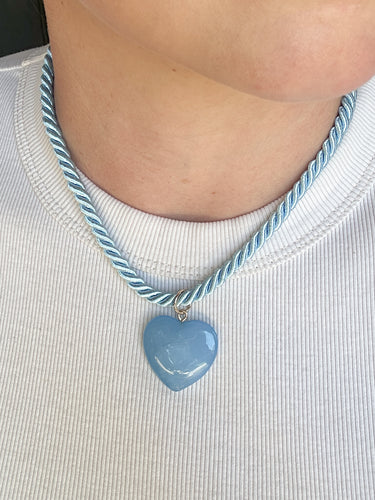 A close-up of a person's neck and chest wearing a light blue heart-shaped pendant necklace with a twisted blue and white rope chain, paired with a white ribbed t-shirt.