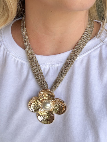 A close-up of a person wearing a chunky gold-tone flower pendant necklace with a textured mesh chain and a central pearl-like bead, paired with a white t-shirt.
