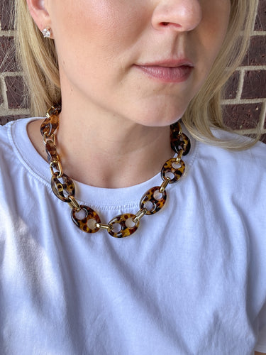 A close-up of a woman wearing a chunky tortoiseshell link necklace with gold-tone metal connectors, paired with a white t-shirt.