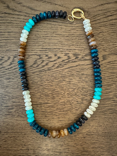 A beaded necklace with a gold clasp, featuring alternating sections of dark blue, black, white, turquoise, and brown beads, displayed on a wooden surface.