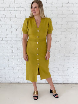 A woman modeling a short-sleeve chartreuse midi dress with a button-down front, standing against a white brick wall.