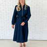 A woman modeling a dark navy blue midi dress with a front zipper, tie waist belt, and long sleeves, standing against a white brick wall.