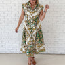 A woman modeling a sleeveless midi dress with a green and gold baroque-style pattern on a cream background, standing against a white brick wall.