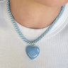 A close-up of a person's neck and chest wearing a light blue heart-shaped pendant necklace with a twisted blue and white rope chain, paired with a white ribbed t-shirt.
