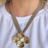 A close-up of a person wearing a chunky gold-tone flower pendant necklace with a textured mesh chain and a central pearl-like bead, paired with a white t-shirt.