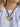 A close-up of a person wearing a chunky gold-tone flower pendant necklace with a textured woven strap and a central pearl-like accent, paired with a white t-shirt.