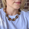 A close-up of a woman wearing a chunky tortoiseshell link necklace with gold-tone metal connectors, paired with a white t-shirt.