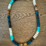 A beaded necklace with a gold clasp, featuring alternating sections of dark blue, black, white, turquoise, and brown beads, displayed on a wooden surface.