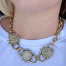 A close-up of a person wearing a gold-toned statement necklace with three coin charms and small clear rhinestone accents, paired with a white top.