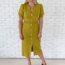 A woman modeling a short-sleeve chartreuse midi dress with a button-down front, standing against a white brick wall.