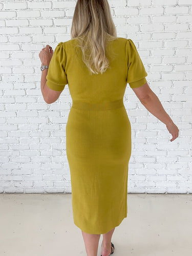 A woman viewed from behind wearing a Chartreuse short-sleeve midi dress with puffed shoulders, standing against a white brick wall.