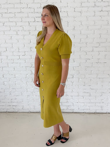 A woman modeling a short-sleeve chartreuse midi dress with puff sleeves and a front button placket, standing against a white brick wall.