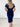 A woman viewed from behind wearing a short-sleeve, bodycon midi dress in dark navy blue, standing against a white brick wall.