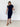 A woman modeling a short-sleeve, bodycon midi dress in dark navy blue, paired with tan block-heel sandals, standing against a white brick wall.