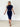 A woman modeling a navy blue short-sleeve bodycon midi dress with a thin brown belt, standing against a white brick wall.