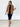 A woman modeling a tan blazer over a navy blue dress, styled with a gold belt, layered necklaces, and tan heeled sandals, standing against a white brick wall.